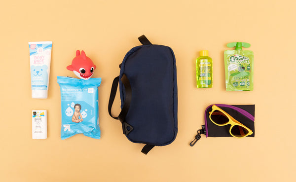 A Navy Parapack Grab Bag with a toddler's items next to it including sunglasses, wipes, sunscreen, and a snack.